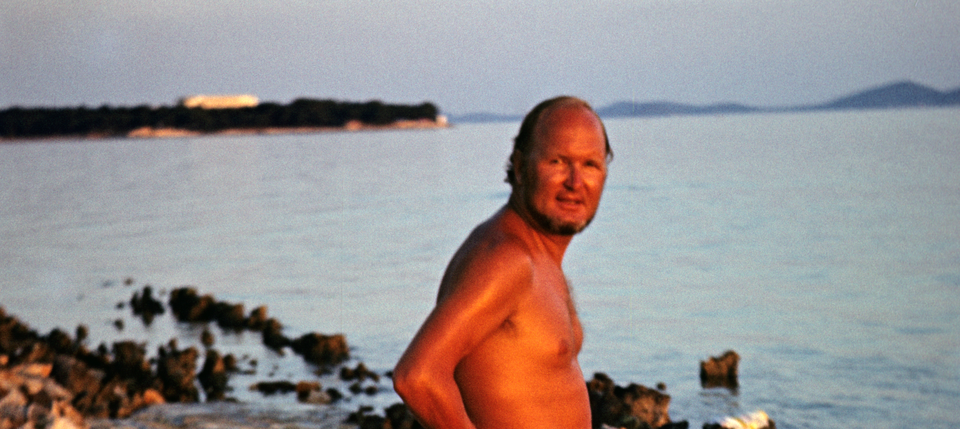 Il padre di Marc è in piedi davanti al mare con la parte superiore del corpo libera e guarda rilassato nella macchina fotografica