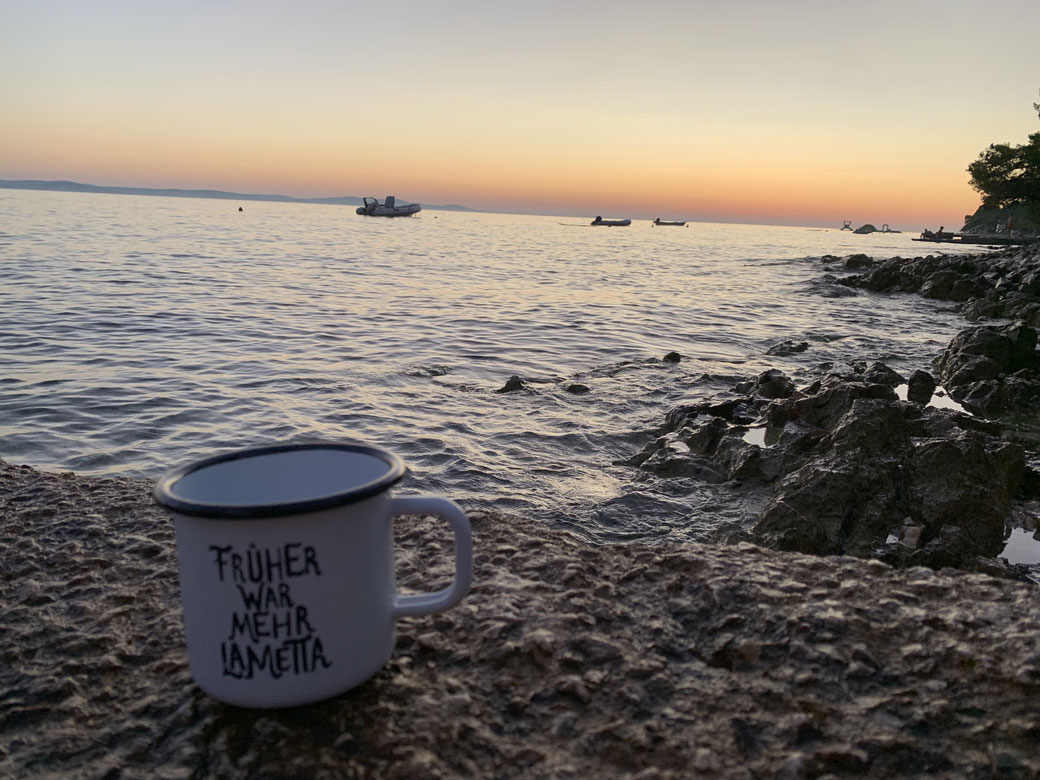 Weiße Emailletasse mit Aufschrift ‚Früher war mehr Lametta’ auf Felsen vor Meer und Sonnenuntergang