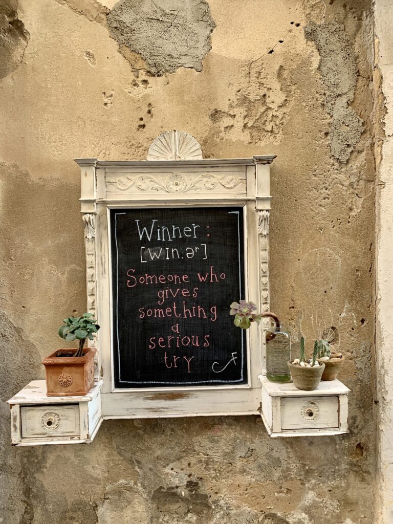 Leistenen plaquette in een steegje in Ortigia met de inscriptie: Winnaar is Iemand die iets serieus probeert